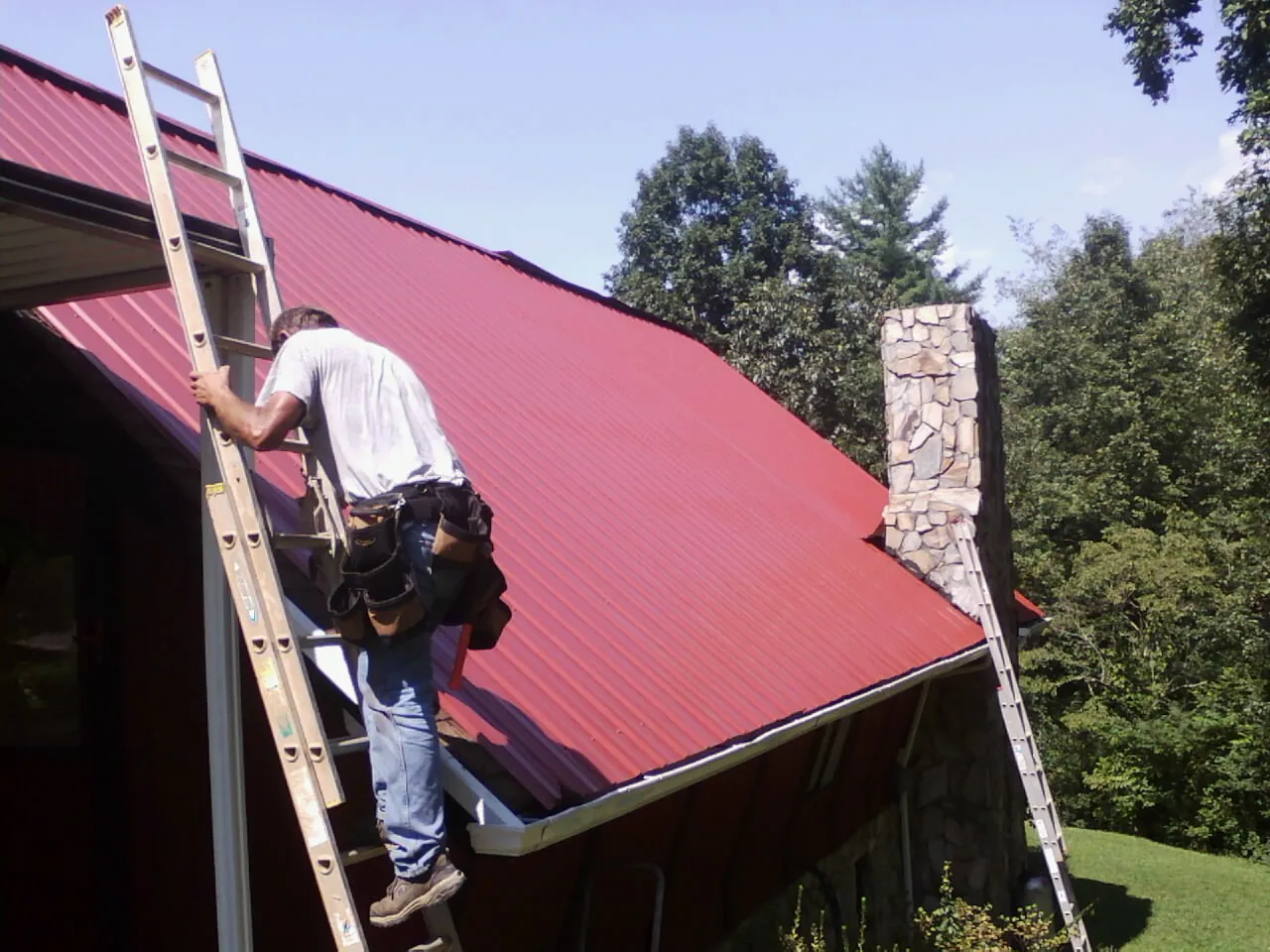 Roofer on a ladder beside a red metal roof and stone chimney — Soffit & Fascia Repair by Roofing Brothers in Frenchville
